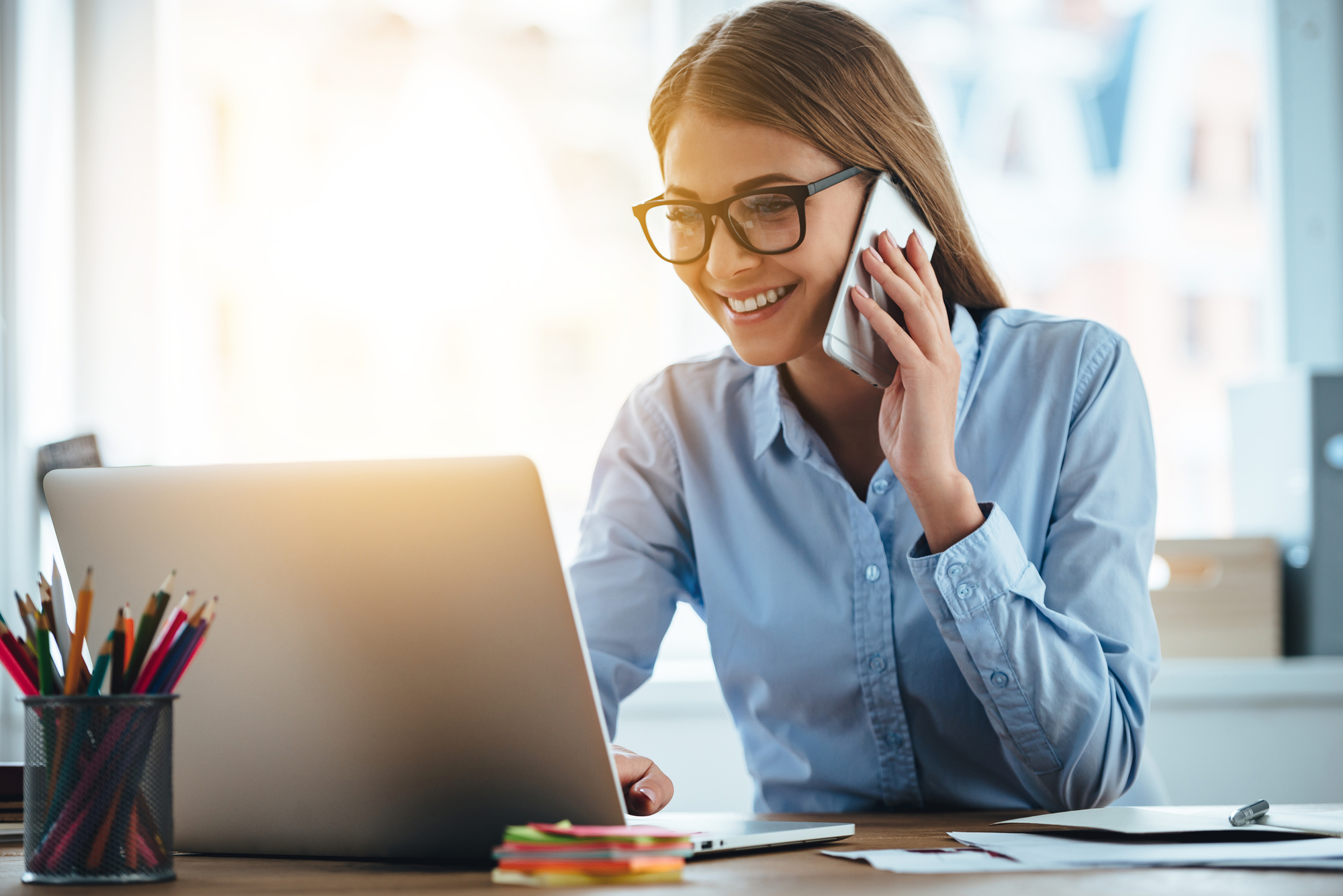 Yes I see your email! Cheerful young beautiful woman in glasses talking on mobile phone and using laptop with smile while sitting at her working place